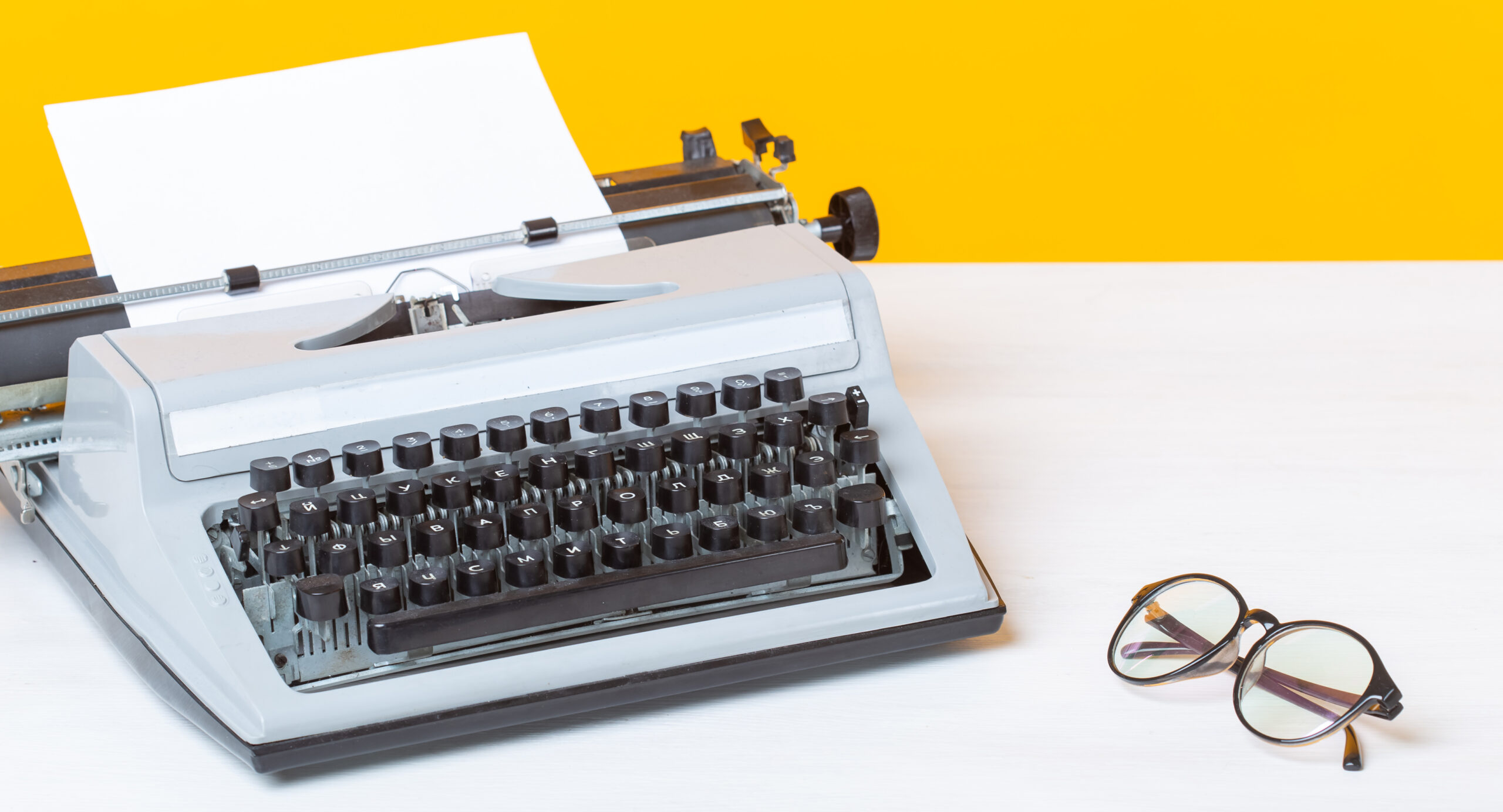 A typewriter with a blank page on a desk alongside some glasses